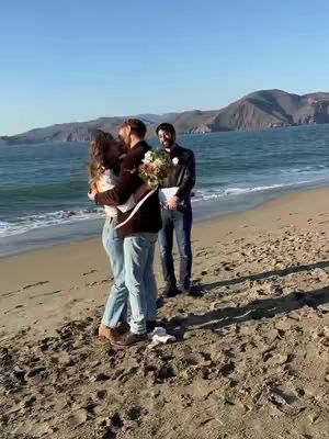 A scenic shot on a beach with two people hugging and kissing, set against a backdrop of ocean and mountains under a clear sky. Another person stands behind them holding flowers and a notebook. The sand is a light tan and the water is a deep blue.