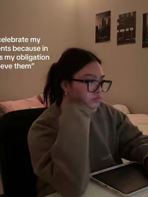 A young woman with dark hair pulled back in a ponytail, wearing glasses and a casual olive-green sweatshirt, is seated indoors. She's looking down, with a neutral expression. Behind her is a sparsely decorated wall and a cozy bed setup, suggesting a relaxed home environment. In front of her sits a tablet and accessories, hinting at a study or work related setup.