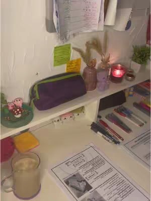 The cover image shows a well-lit desk area with a variety of items indicative of a study or work space. At the bottom left, a clear glass mug filled with a light-colored beverage sits on the desk. To the right, a printed paper lies, and in front of it, pens are arranged on a shelf. A purple pencil bag and an assortment of decorative items like small vases and a candle add character to the space. The background of the composition features white walls and some sticky notes, as well as notes attached to the shelf backing, contributing to the image's organized and inviting feel.