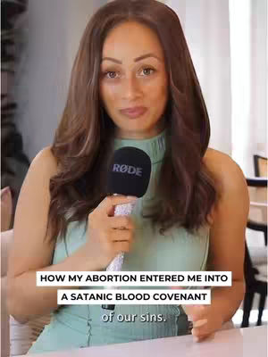 A woman with long, wavy brown hair is holding a microphone in front of her. She is wearing a pale green, sleeveless top. The background is blurred, but appears to be a room with simple decor and light-colored walls. Text overlays appear at the bottom of the image.