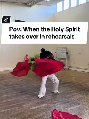 The image is a vertical shot showing a dance studio during a rehearsal. The focal point is a woman with red fabric in her hands, in the foreground, actively dancing. The room has wooden floors, with light coming through the windows.