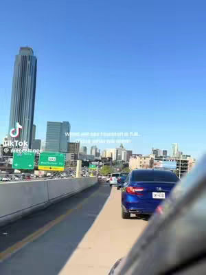 A view from inside a car traveling on a highway. The highway is filled with cars. In the background, there are multiple skyscrapers and buildings. The sky is clear blue. The foreground consist of the inside elements of a car with a blue car on the right side.