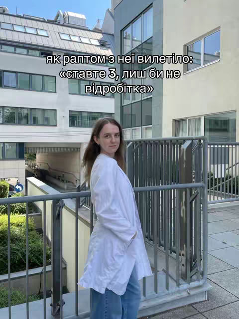 A woman in a white lab coat stands on a balcony with a metal railing, overlooking a building with large windows. The sky is blue, and the overall color palette is bright and natural. Text overlays are present on the image.
