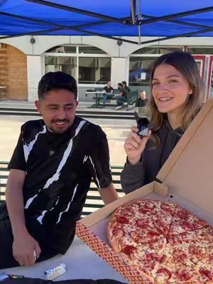 Two people pose for a photo outdoors. A man is seated, while a woman stands holding a pizza box open to reveal a pepperoni pizza. The setting appears to be in front of a building with a blue canopy overhead.