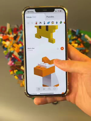 The cover image shows a person holding a phone displaying an app interface. The phone is surrounded by scattered colorful building blocks. On the phone screen, two toy block model designs are visible, with one highlighted by a finger touching the screen. The background is a mix of colorful blocks and a plain surface.