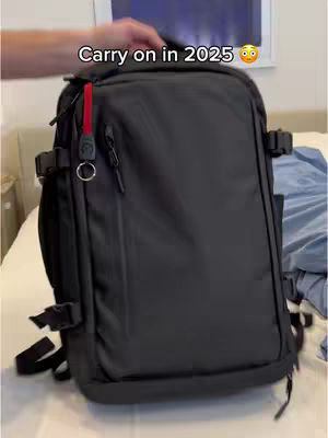 The image displays a large black backpack, centered in the frame. The backpack has several pockets, straps, and buckles. A hand is visible at the top of the bag, presumably holding it. The background includes a bed with a white cover and a light-colored bedside table. The overall composition highlights the backpack's features and design.