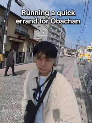 A selfie-style shot of a young man standing on a city street in Japan. He is the central focus, looking directly at the camera with a neutral expression. The background shows a typical Japanese residential area with buildings and streets. The sky is clear, suggesting a sunny day. The foreground shows the street and some plants.