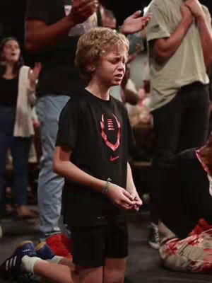 A young boy kneels on the floor, eyes closed in prayer or worship. Surrounding him are other people, some with their hands raised as well. The lighting suggests a church setting, with soft, warm tones.