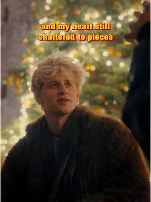 A young man with messy blonde hair is gazing upwards with a slightly concerned expression. He is wearing a dark coat or jacket. Behind him is a blurry background featuring a Christmas tree with warm, yellow lights.
