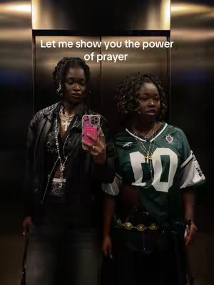 Two young women stand side-by-side in an elevator, both dressed in stylish attire. The woman on the left is wearing a black leather jacket and holding a phone, while the woman on the right is in a green jersey. The elevator interior is metallic with a bright light giving the scene a moderate level of illumination