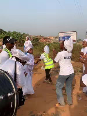 A group of people in white garments are dancing on a dirt path. Some have hats and vests. The background has buildings and trees.