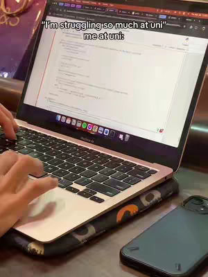 A person is working on a laptop, the screen displaying code, with a phone next to it on a desk. A text overlay stating "I'm struggling so much at uni" above "me at uni:".