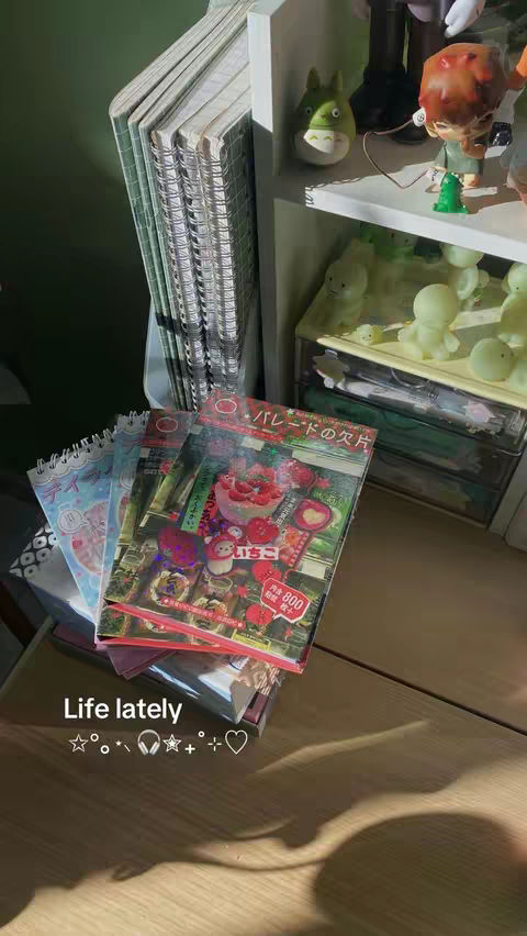The image is of a desk with several layered notebooks and books, with a Japanese-style book cover. Various knick-knacks are on a top shelf. The lighting is natural, and the colors are warm.