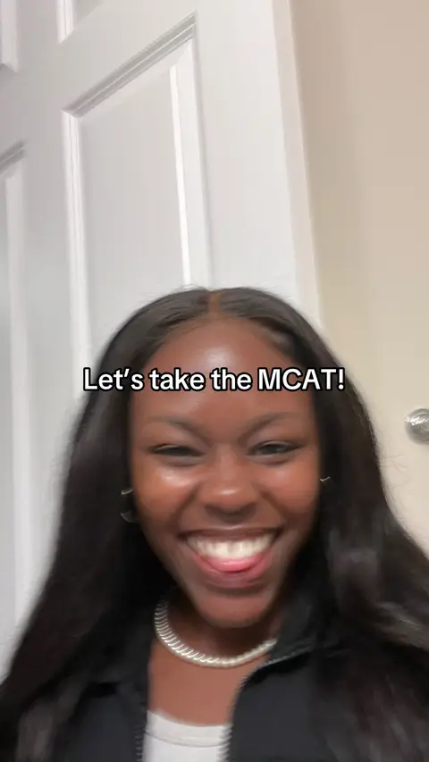 A close-up shot shows a smiling Black woman. Her dark hair is long and straight, framing her face. She wears a dark jacket and a silver necklace. Text overlayed on the image reads "Let's take the MCAT!"