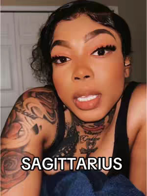 A selfie-style shot of a woman with dark hair, makeup, and tattoos. The image is dominated by her face and upper body, and a text overlay at the bottom reads 'SAGITTARIUS'.