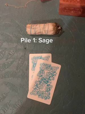 The image shows a close-up of a gray-green table surface holding a sage bundle, and two tarot cards facing away from the viewer. The sage bundle is beige and wrapped in a green string, with the tip charred black. The tarot cards are pale with blue floral designs on the back. On the upper right of the image, a wooden object with golden color can be seen. The image is lit with a flat, natural light.