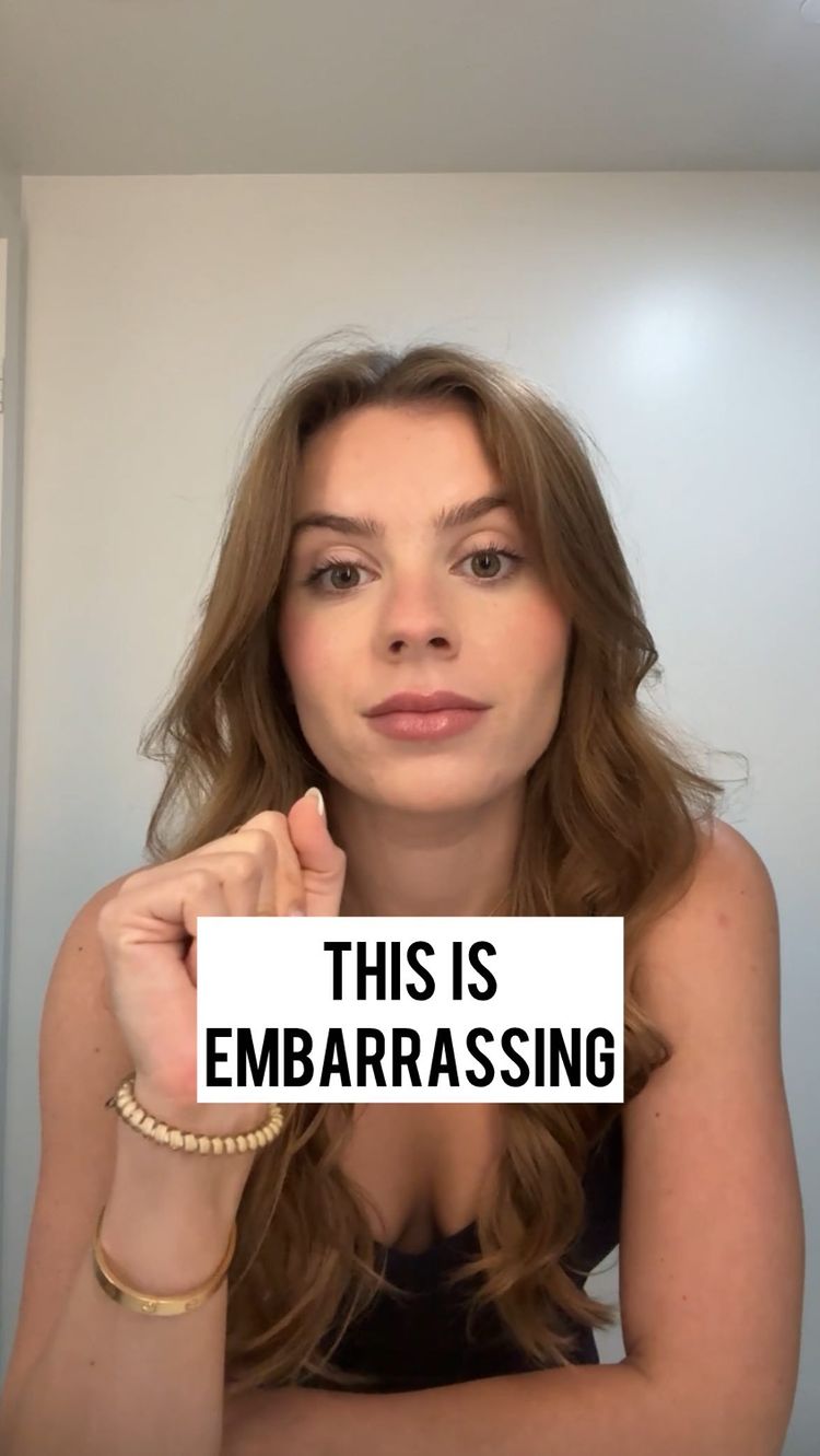 A woman with long, wavy brown hair is centered in the frame. She is looking directly at the viewer, resting her right hand near her face, close to her lips. A white rectangle with the text 'THIS IS EMBARRASSING' in black, bold font is overlaid on her chest. She wears a black top and a gold bracelet on her left wrist.