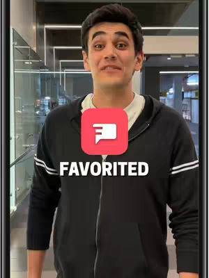 A man smiles at the camera, with a red speech bubble icon and the word "FAVORITED" overlaid on his torso. He is wearing a black zip-up hoodie over a white t-shirt, standing in what appears to be a hallway with glass paneling.