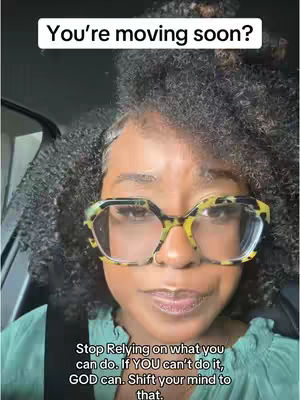 A close-up shot of a Black woman inside a car, with dark, curly hair framing her face. She wears large, thick-rimmed glasses with a tortoiseshell pattern. The background shows the interior of a vehicle with the car door frame visible. The woman has a gentle expression and appears to be speaking or reacting to something.
