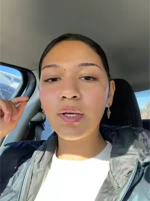 A woman with dark hair is sitting in a car. She is looking at the camera and has her mouth slightly open as if she is about to speak. The background suggests the car's interior with a window visible.