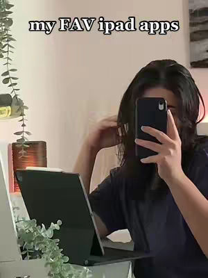 A selfie-style shot featuring a young woman holding her phone up to her face, partially obscuring it. She's also holding an iPad open with a keyboard attachment. The foreground includes foliage like plants, suggesting a well-lit indoor setting. The background is somewhat blurred, with hints of a neutral wall and a decorative element.