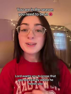 A young woman with long brown hair and glasses is centered in the frame, facing the camera and speaking. Text overlays appear above and below her, conveying a religious message. The background shows a room with soft lighting and what appears to be Christmas lights.