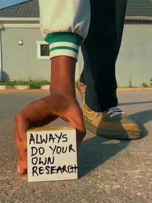 A person's hand holds a small, yellow cube with the text "ALWAYS DO YOUR OWN RESEARCH" in black, against a blurred background that appears to be a street scene. The person's arm, wearing a white jacket with green accents, is visible. The person is also wearing dark pants and a light-colored sneaker.