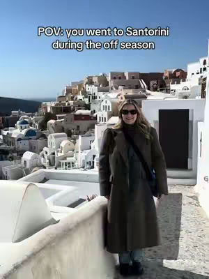 A woman in a long coat and sunglasses poses on a white stone walkway overlooking a picturesque Greek village with white buildings and blue accents on a bright sunny day. The sky is a clear blue.