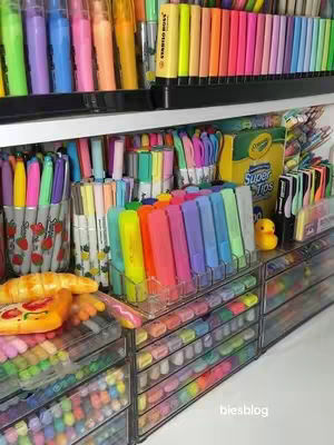 A colorful, neatly organized display of stationery items fill the frame. Rows of multi-colored pens, markers, and pencils are arranged on shelves and in transparent drawers, offering a visually appealing and organized aesthetic. The composition highlights the diversity of colors and types of stationery.