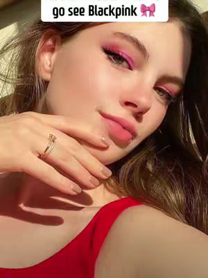 A selfie-style close-up that features a woman with long brown hair, pink eyeshadow, and a red top pointing at her face. The background is blurred. A gold ring is on her finger.