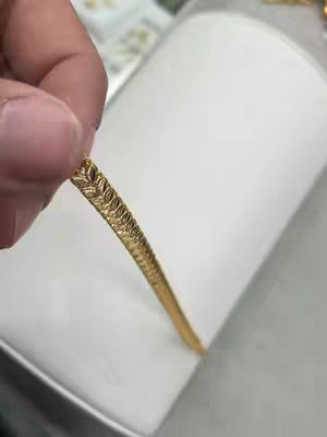 A close-up shot of a hand holding a gold bracelet. The bracelet is in focus, showcasing a detailed leaf-like pattern. The background is blurry, showing a white surface.