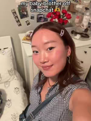 A selfie-style shot featuring a young woman positioned in the foreground. She has brown hair, styled with a side part, and a few strands are pulled back with a white barrette. She's wearing a black and white checkered shirt, and a dark strap from a bag is visible across her chest. Behind her, a bed with white pillows is partially visible, and a white dresser is set up in a dimly lit room. On the wall behind her, there are various pictures and decorations.