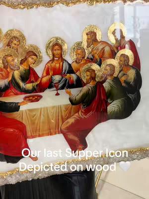 The image displays an artistic representation of the Last Supper, rendered in a style reminiscent of religious icons. Jesus and his disciples are gathered around a table, with Jesus seated centrally and extending his hand towards a chalice. The scene is richly colored, featuring gold leaf accents around the figures' halos. The composition is framed within an irregularly shaped border. The overall effect is one of reverence and artistic craftsmanship.