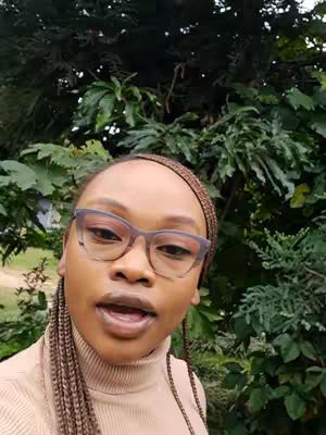 A woman with brown skin stands close to the camera, her head and shoulders visible. She has long, dark brown braids. She is wearing glasses while talking. Behind her, green leaves are densely packed, creating a natural backdrop.
