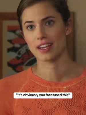 A woman with fair skin and light brown hair is looking slightly to the right and speaking or singing. She is wearing an orange top, and there is a framed picture on the wall behind her.