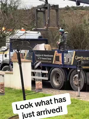 The image shows a man standing on a truck bed in a rural environment. The truck is a large, dark-colored vehicle with signage indicating it is a supplier of natural stone. A mechanical arm is lifting what appears to be a glass object into position. The man is dressed in a jacket and hat. The background includes a mix of trees and bushes, with a calm, overcast sky suggesting a cool, cloudy day. A text overlay in a white box at the bottom of the image says 'Look what has just arrived!'