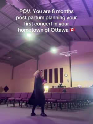 A woman in a long black coat and dark hair with a church interior and purple chairs. Text overlay at the top says: “POV: You are 8 months post partum planning your first concert in your hometown of Ottawa 🇨🇦”.