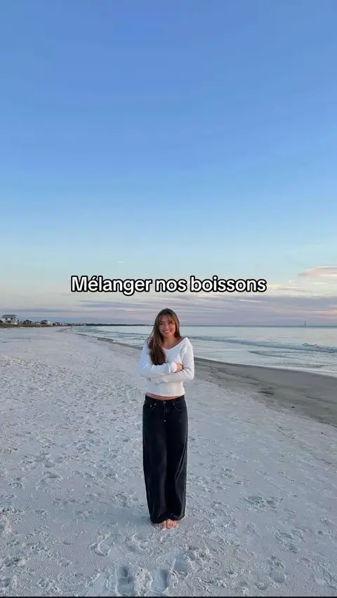 A serene scene unfolds on a sandy beach under a vast, cloudless sky. A woman with long, brown hair stands barefoot on the white sand, facing the camera with arms crossed. The woman is wearing a white sweater and dark colored pants. The horizon line, where the ocean meets the sky, is visible in the background.
