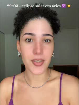 A woman with curly dark hair faces the camera in a close-up shot. She wears silver hoop earrings, a silver necklace, and a purple spaghetti strap top. The background is blurred, suggesting an interior setting. Text overlayed at the top reads, "29/03 - eclipse solar em áries" (solar eclipse in aries).