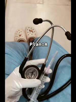 The image shows a person's lap and lower body in blue scrubs. In their gloved hand, they hold a stethoscope, and a syringe is being held in their had. In the background at the top of the image are visible white Crocs.
