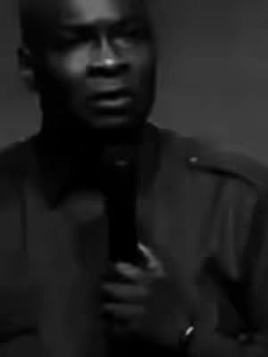 A grayscale image showing a man in a dark suit jacket, holding a microphone. The focus is on his face, angled slightly up, with his mouth open as if speaking. A plain background contrasts with the darker tones of his attire and skin.