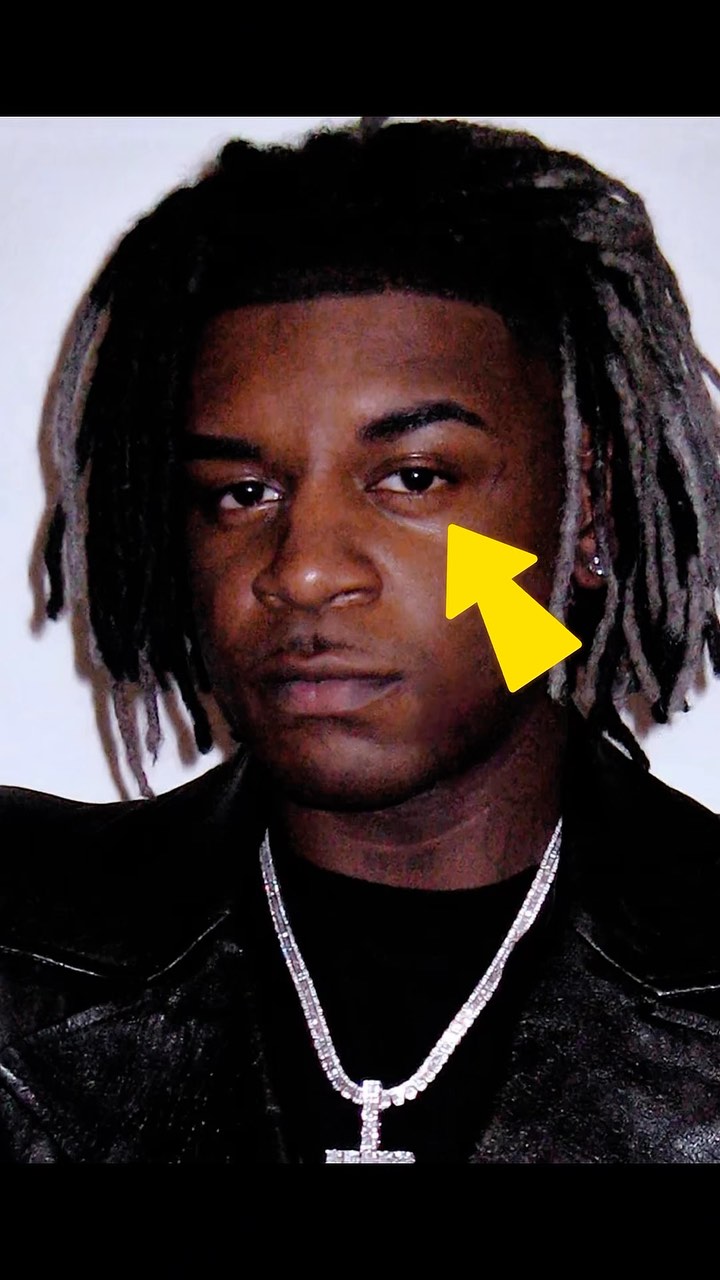 Close-up shot of a man with a stern expression, a black leather jacket, and a diamond necklace; a yellow arrow points to a scar on his face. His hair is styled with grey-tipped dreadlocks. The background is a plain white.