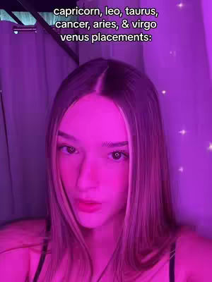 A selfie-style shot of a woman with long hair, framed from the chest up. Her face is lit with pink light, and a text overlay reads the names of several zodiac signs. The background is blurred, suggesting a soft, ambient setting, with a hint of some kind of curtain or backdrop.