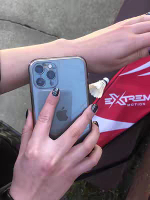 A close-up shot shows a hand holding a smartphone. The hand has dark nail polish, and the phone has a clear case. A badminton shuttlecock sits nearby, and a red and white bag is partially visible.