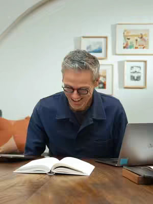 A man with short gray hair and glasses is sitting at a wooden table, looking down with a smile. An open book is in front of him, and a laptop and a phone are to his right. The background includes framed art pieces and a section of a wall.