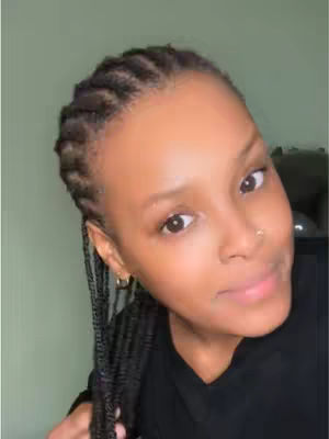 A selfie-style shot of a woman with dark skin and braids. She's looking to the right with a slight smile. Her eyebrows are well-defined, and she has a small nose stud. She's wearing a black top, and the background is a simple, muted green.