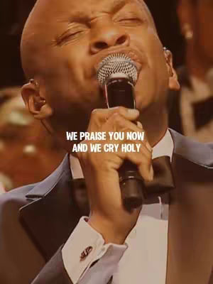 A close-up shot of a bald Black man singing into a microphone with visible emotion. The background is blurred, suggesting a performance setting.