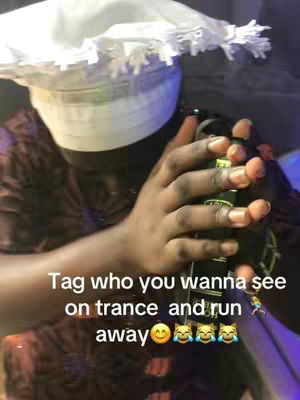 A person of color is shown in a close-up shot, holding a microphone. They are wearing a white, ruffled head covering and a patterned brown shirt, with their hands prominently displayed. Text on the image encourages viewers to tag someone "on trance" with emojis. The lighting is dim, casting shadows, and the composition is a mix of the person and the surrounding environment.