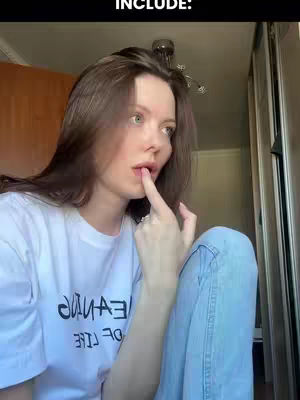 A young woman with long brown hair and fair skin looks off to the side with a somewhat pensive expression. She is wearing a white t-shirt and light blue jeans. She is indoors, suggesting a casual setting. She is touching her bottom lip with her index finger.