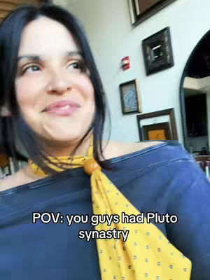 A woman with dark hair poses for a selfie, wearing a dark blue top and a yellow scarf. The background includes artwork and a mirror.